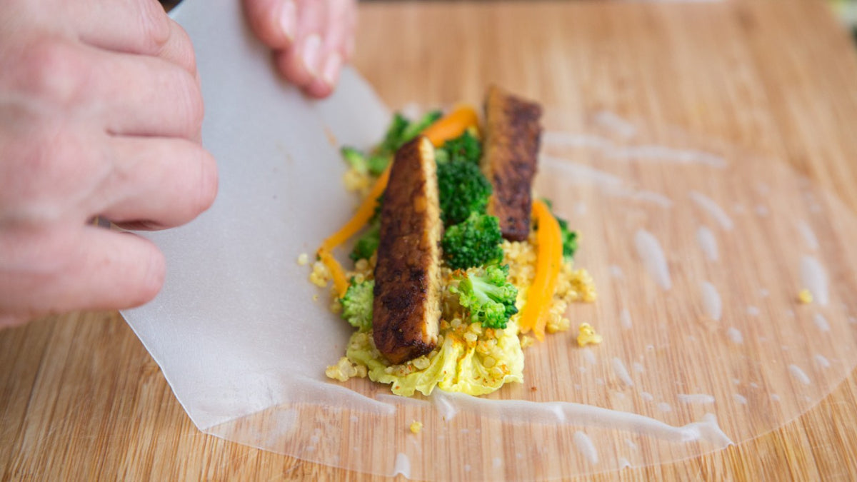 Auf einer Holzunterlage werden mit den Händen frische Reispapier-Wraps mit Gemüse und gegrilltem Tempeh gerollt.