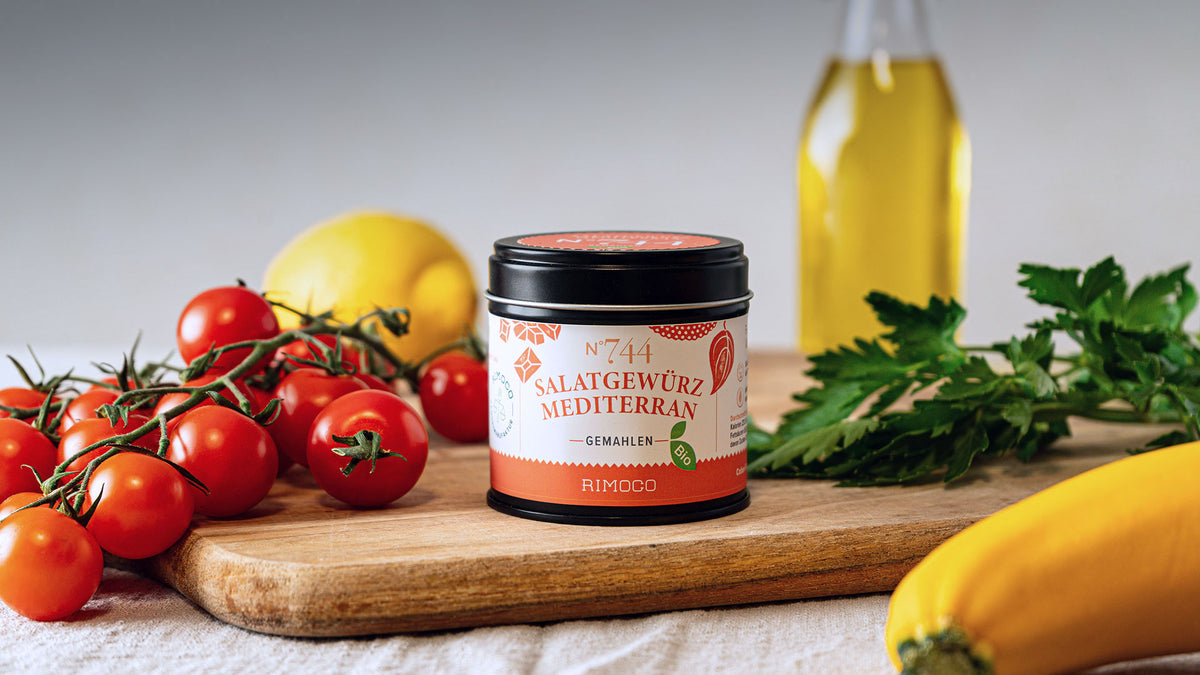 Mediterrane Gewürzdose auf einem Brett mit Tomaten, frischen Kräutern, Zitrone und Olivenöl.