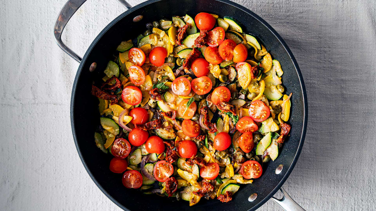 Eine Pfanne mit gebratenem Gemüse und Kirschtomaten auf einem hellen, rustikalen Untergrund.
