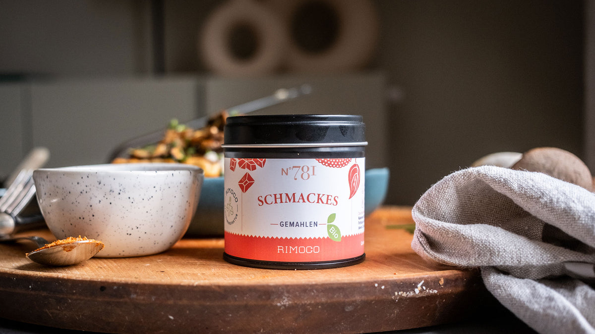 Eine Schmackes-Dose auf einem Holzbrett, daneben eine Schale, ein Löffel und ein Tuch.