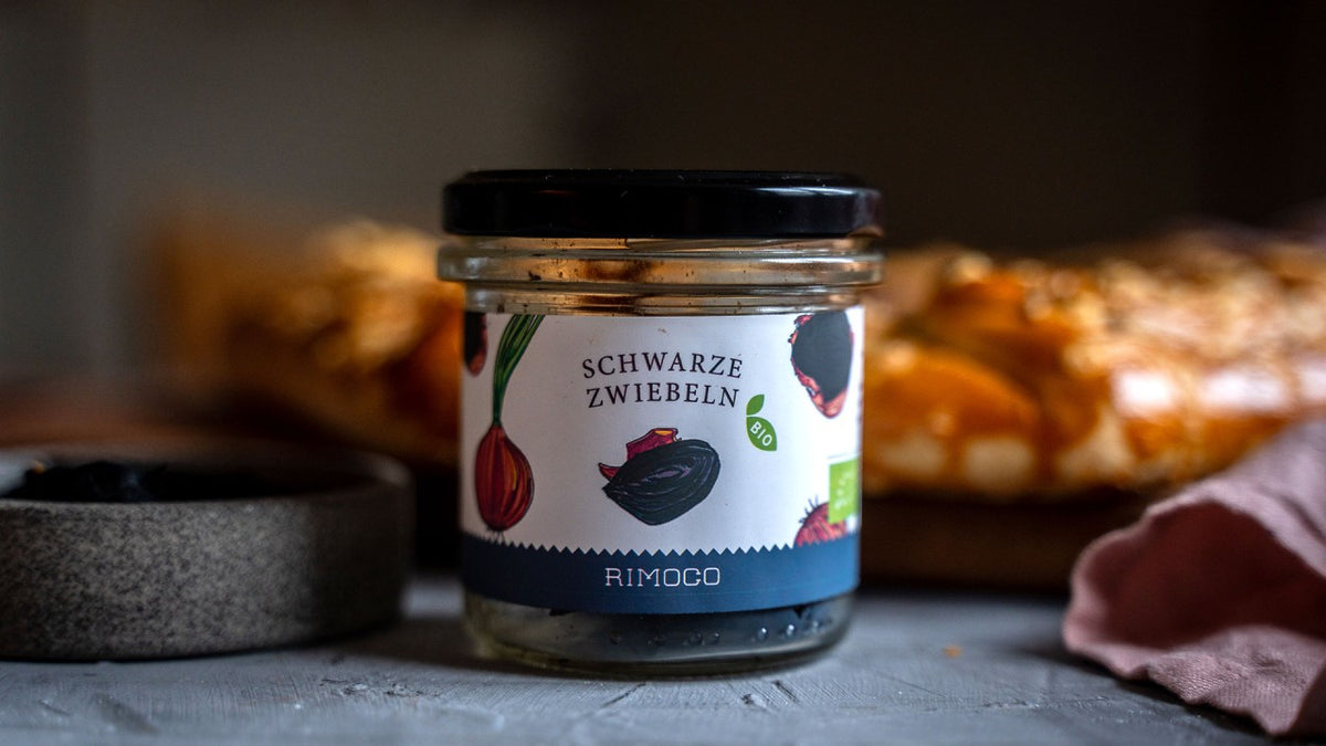 Auf einem Tisch steht ein Glas mit der Aufschrift „Schwarze Zwiebeln“ neben frischem Brot und einer kleinen Schale.