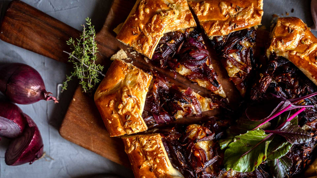 Herzhafte Tarte mit fermentierten schwarzen Zwiebeln, frischen Kräutern und Salat auf Holzbrett.