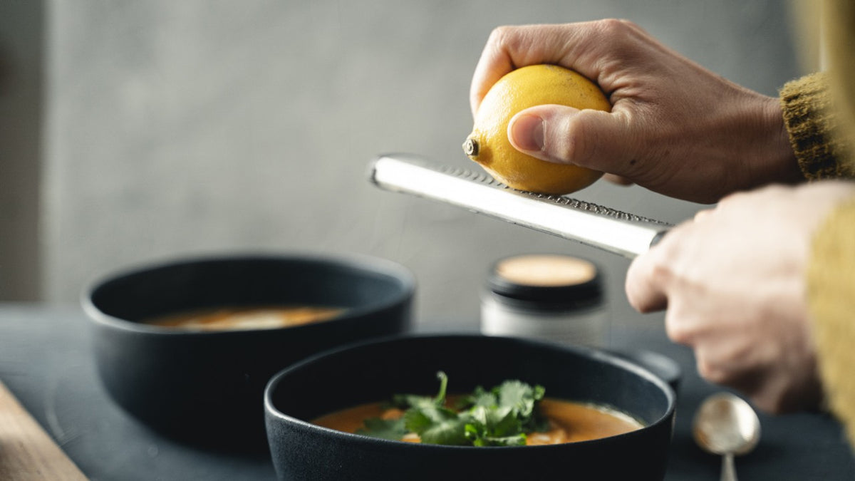 Im Vordergrund steht eine Kräutersuppe, darüber reibt jemand Zitronenschale, im Hintergrund steht eine zweite Schüssel.