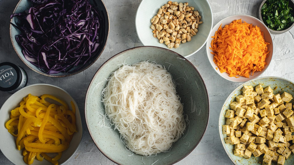 Schüsseln mit Reisnudeln, Tofu, Karottenstreifen, Erdnüssen, gelber Paprika, Kohl und frischen Kräutern.
