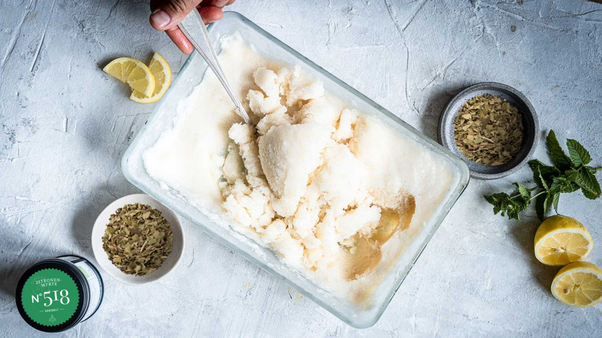 Eine Hand rührt Zitronensorbet in einer Glasschale um, daneben liegen Zitronenscheiben und eine Schälchen mit Fenchelsamen.