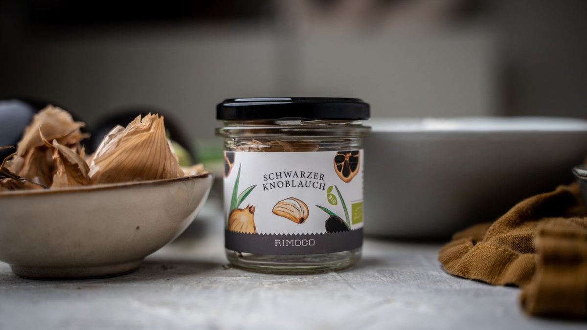 Auf einem Tisch steht ein Glas mit der Aufschrift „Schwarzer Knoblauch“ neben einer Schale und Knoblauchschalen.