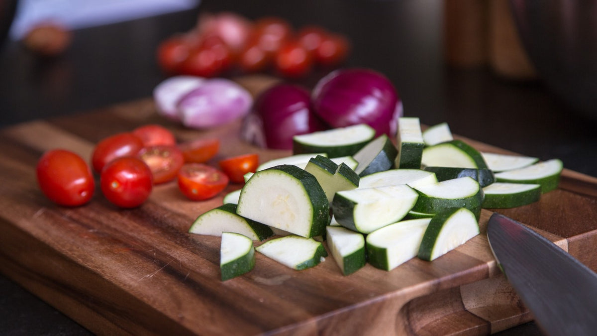 Geschnittene Zucchini, Kirschtomaten und rote Zwiebeln auf einem Holzbrett mit einem scharfen Messer.