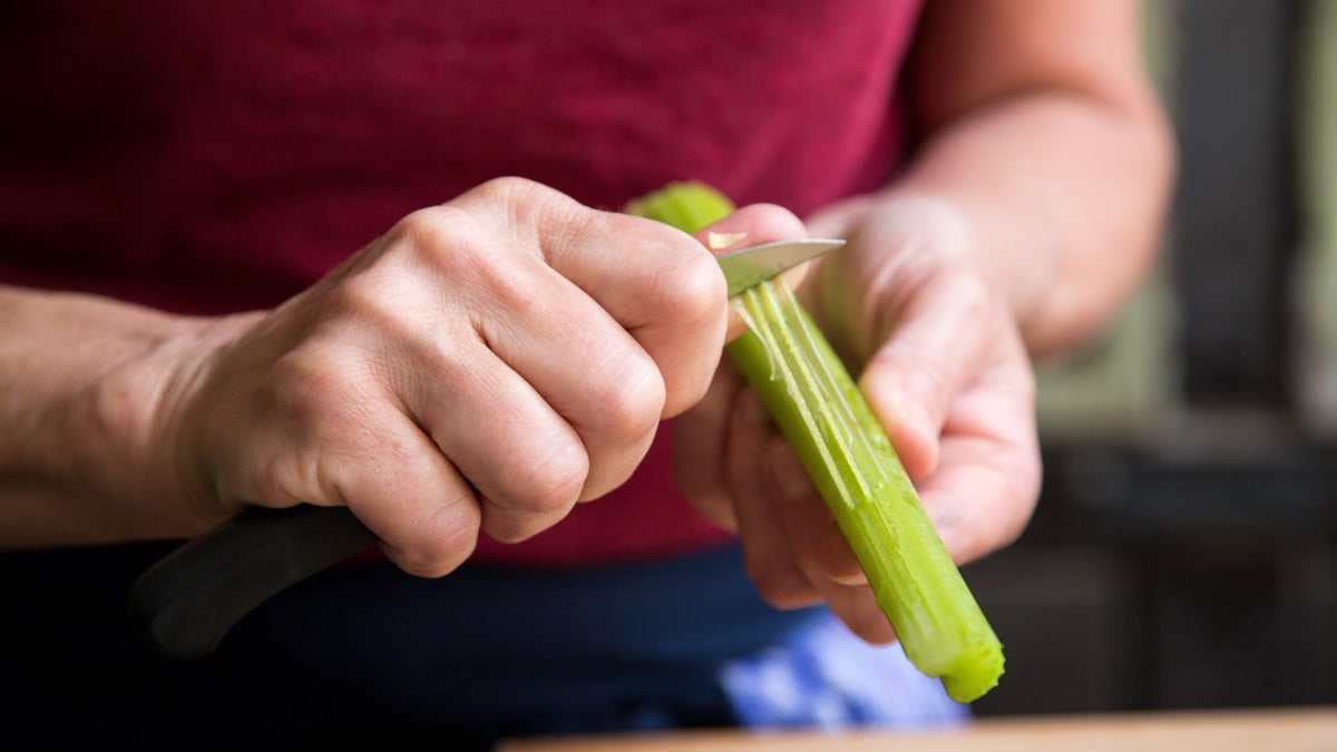Hände schälen einen frischen Selleriestängel mit einem kleinen Messer, rote Kleidung, Nahaufnahme.
