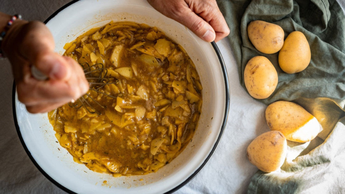 Mit den Händen werden Kartoffelscheiben in gelber Soße vermengt, ganze Kartoffeln liegen auf einem Tuch.