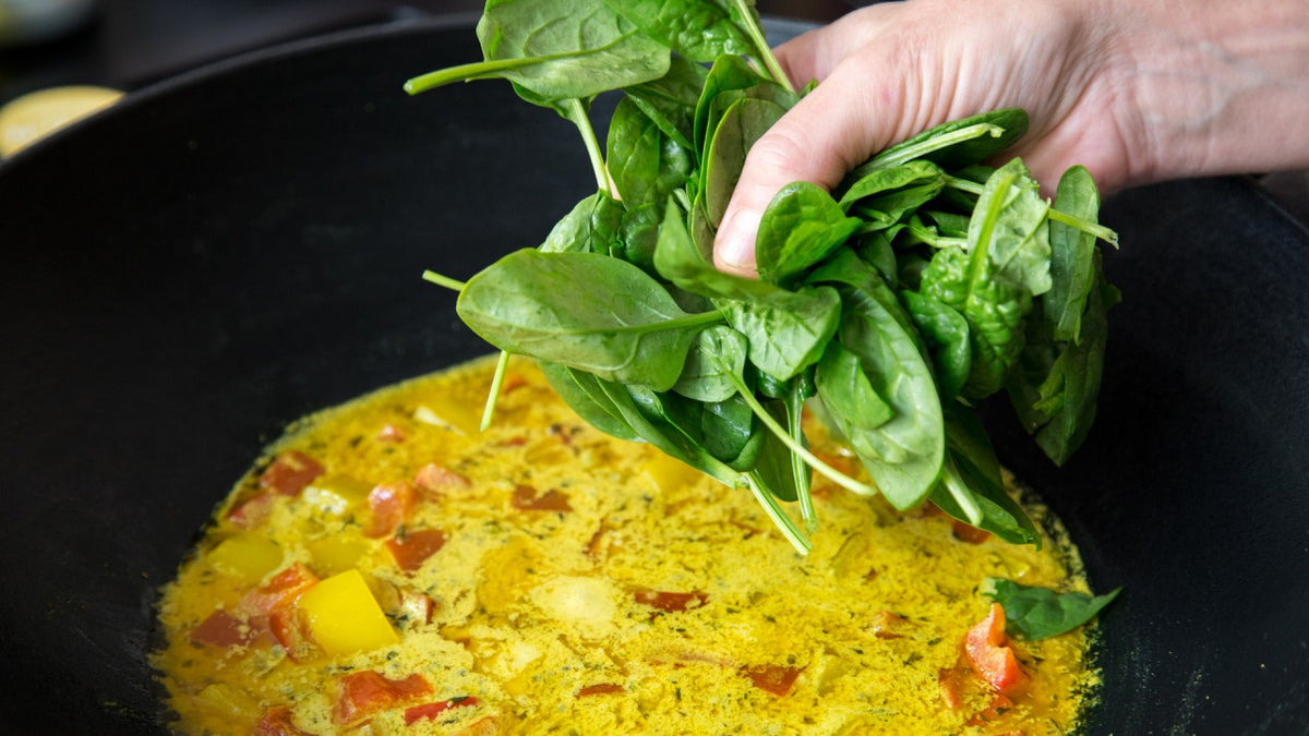 Gelbes Curry mit frischem Spinat in einer schwarzen Pfanne; der Spinat wird gerade hinzugefügt.