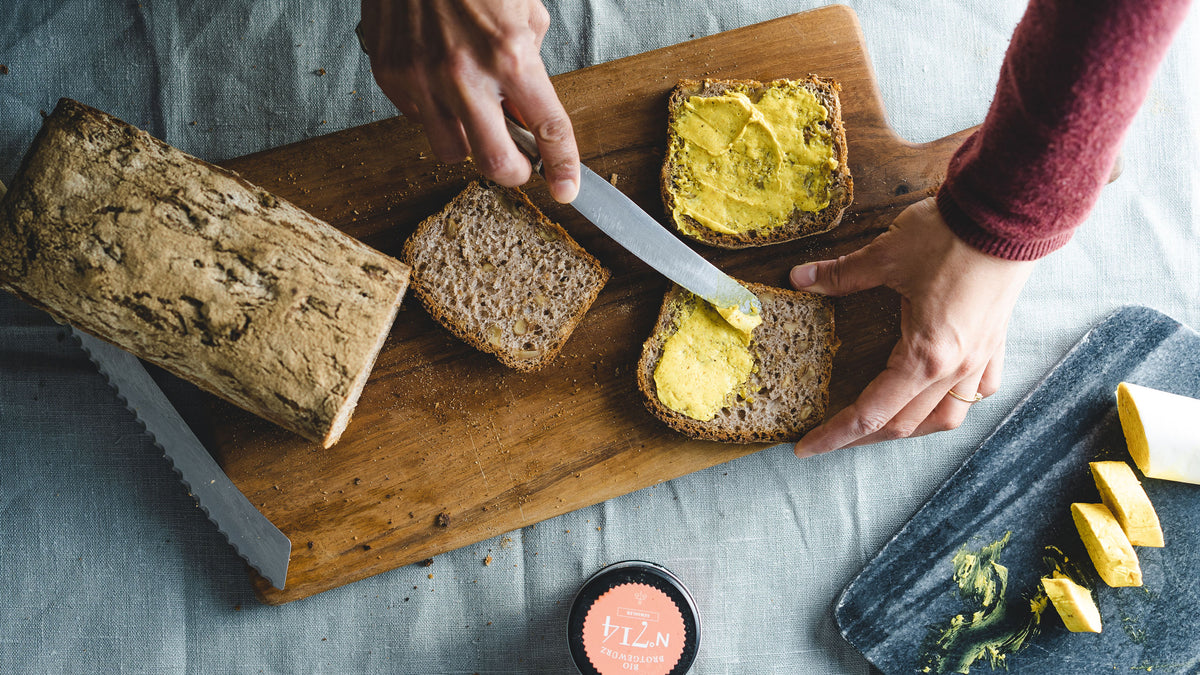 Hände streichen gelbe Butter auf Brot, daneben ein magnetisches Gewürzregal mit 12 sichtbaren Gewürzen.