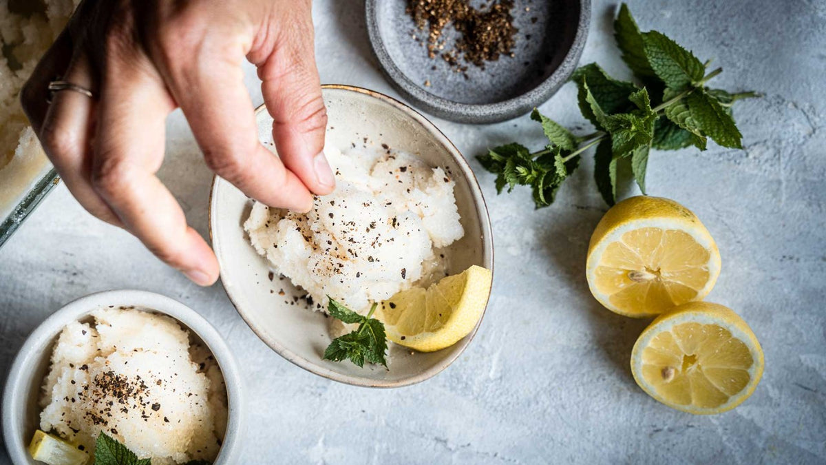 Eine Hand streut Pfeffer über Zitronensorbet, garniert mit Minze und frischen Zitronenscheiben.