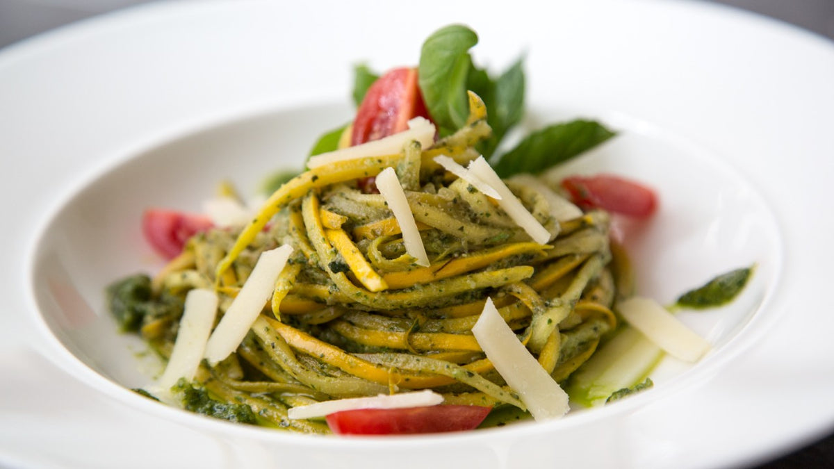 Tagliatelle mit grünem Pesto, garniert mit Käsespänen, Basilikumblättern und Tomatenscheiben.